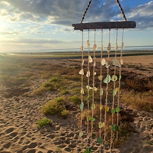 Peut inclure: Un mobile suspendu composé d'un morceau de bois flotté horizontal retenu par une corde torsadée. Plusieurs brins verticaux de ficelle sont fixés au bois, chacun orné de divers morceaux de verre de mer translucide dans des tons verts et blancs.