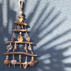 Peut inclure: Une décoration murale artisanale en forme de sapin de Noël, composée de bois flotté, de coquillages et de perles en bois. Les tons naturels des matériaux contrastent avec le mur bleu. La suspension est assurée par une boucle tressée.