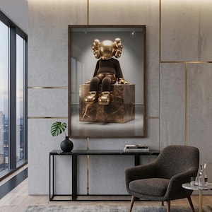 May include: A framed wall art print featuring a golden KAWS character with X-shaped eyes sitting on a brown marble cube. The figure has a brown torso, golden hands, and golden shoes, displayed in a modern room with a dark console and armchair.