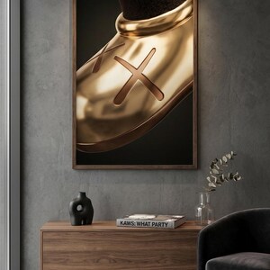 May include: A framed wall art print featuring a close-up of a shiny gold-toned shoe with a prominent X symbol on the side. The artwork is displayed in a dark wood frame, showcasing a smooth, reflective metallic surface against a dark background.