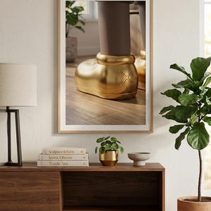 May include: A framed artwork featuring a close-up of a golden boot sculpture. The boot has a rounded shape with an X design on the side. The artwork is displayed on a wooden cabinet with books and a small plant.