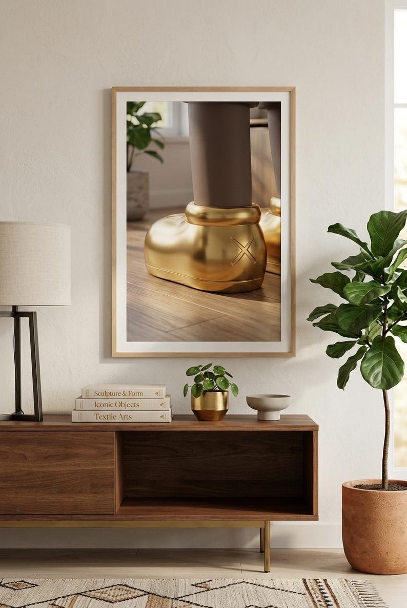 May include: A framed artwork featuring a close-up of a golden boot sculpture. The boot has a rounded shape with an X design on the side. The artwork is displayed on a wooden cabinet with books and a small plant.