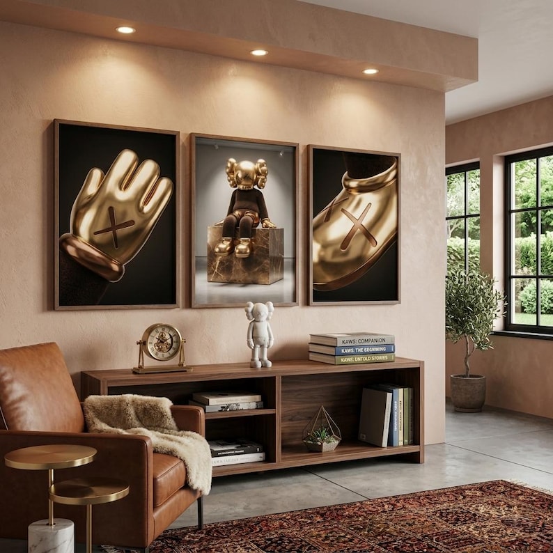 May include: Three framed wall art prints featuring gold-colored cartoon-style hands and a seated figure with X-shaped eyes. Below, a white figurine stands on a wooden console table next to a gold clock and a stack of books titled KAWS.