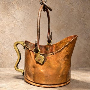 May include: A copper coal scuttle with a hammered texture, featuring a curved brass handle on the side and a tall, dark iron swing handle attached to the rim with metal brackets. The vessel has a wide, angled opening for pouring.
