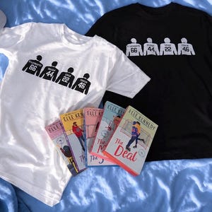 May include: Two t-shirts, one white and one black, each with a graphic of four figures with jersey numbers. The white shirt has the numbers 66, 44, 22, and 46. The black shirt has the same numbers. Several copies of the book "The Deal" by Elle Kennedy are also visible.