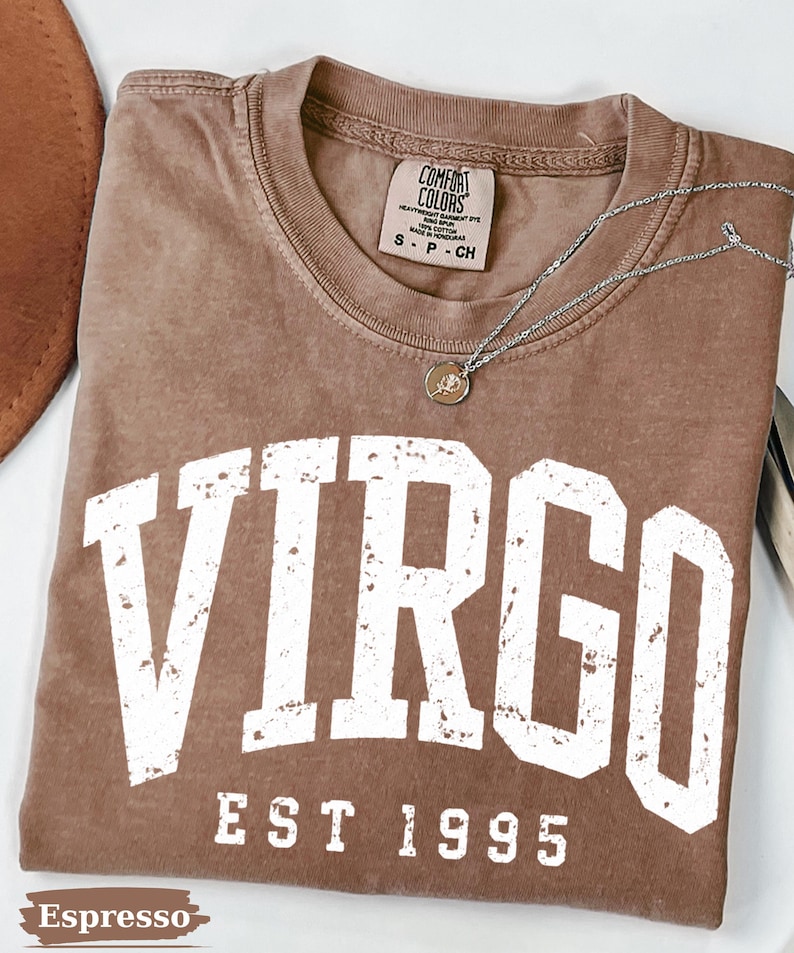 May include: A brown short-sleeved t-shirt with a distressed white graphic that reads VIRGO EST 1995. The shirt features a Comfort Colors brand label at the inner neckline indicating size S, and a silver-toned chain necklace with a small round pendant.