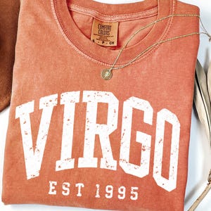 May include: A terracotta-colored short-sleeve t-shirt with a crew neckline. The shirt features the white text VIRGO and EST 1995 in a distressed collegiate font. A gold-toned necklace with a small circular pendant rests on the fabric.