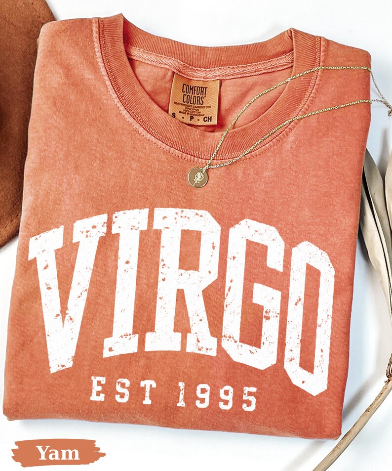 May include: A terracotta-colored short-sleeve t-shirt with a crew neckline. The shirt features the white text VIRGO and EST 1995 in a distressed collegiate font. A gold-toned necklace with a small circular pendant rests on the fabric.