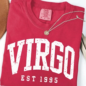 May include: A red short-sleeved t-shirt featuring the word VIRGO in large, distressed white capital letters, with the text EST 1995 printed below in a smaller font. The shirt has a Comfort Colors brand label inside the collar.