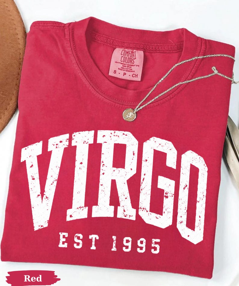 May include: A red short-sleeved t-shirt featuring the word VIRGO in large, distressed white capital letters, with the text EST 1995 printed below in a smaller font. The shirt has a Comfort Colors brand label inside the collar.