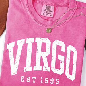 May include: A pink short-sleeve t-shirt featuring the white distressed text VIRGO EST 1995 printed across the chest. The shirt has a Comfort Colors brand label at the inner neckline and is paired with a thin silver-toned necklace with a small circular pendant.