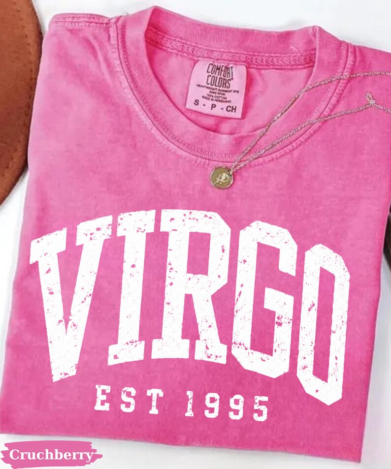 May include: A pink short-sleeve t-shirt featuring the white distressed text VIRGO EST 1995 printed across the chest. The shirt has a Comfort Colors brand label at the inner neckline and is paired with a thin silver-toned necklace with a small circular pendant.