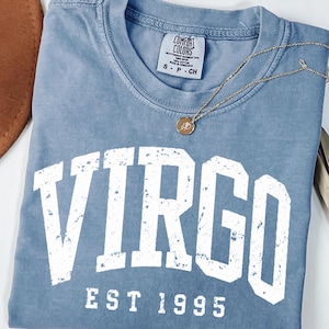 May include: A blue t-shirt with a distressed white graphic that reads VIRGO EST 1995. The shirt features a Comfort Colors brand label at the inner neckline indicating size S, and a thin gold-toned chain necklace with a small circular pendant rests on top.