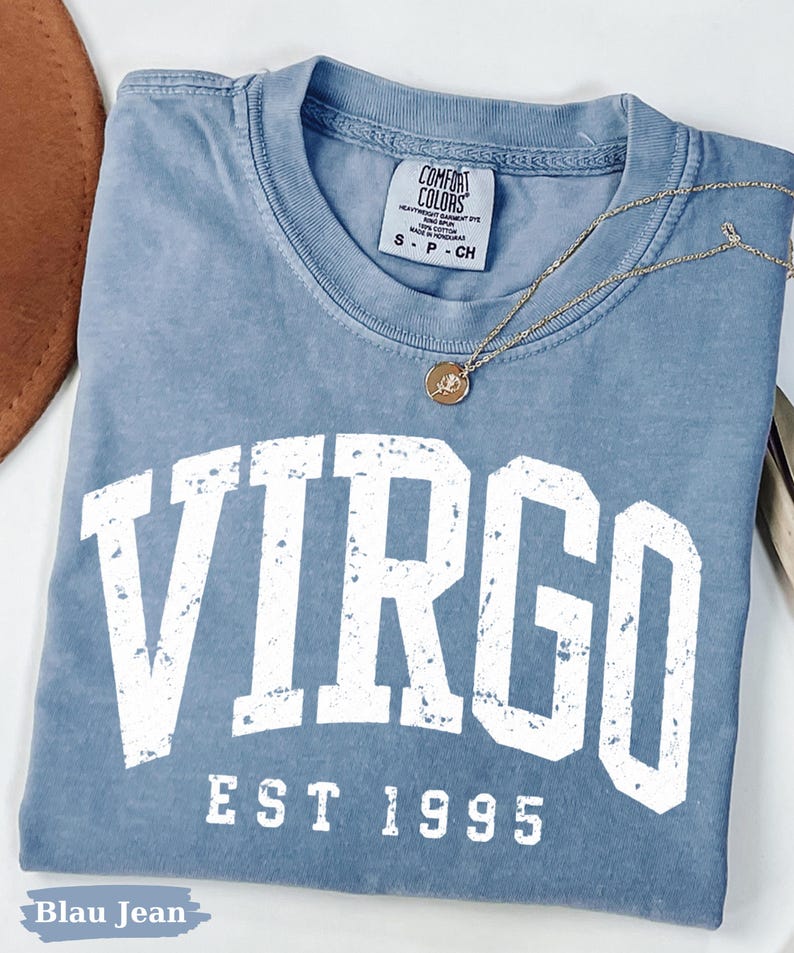 May include: A blue t-shirt with a distressed white graphic that reads VIRGO EST 1995. The shirt features a Comfort Colors brand label at the inner neckline indicating size S, and a thin gold-toned chain necklace with a small circular pendant rests on top.