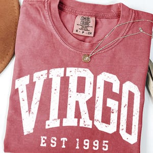 May include: A crimson short-sleeved t-shirt featuring the white distressed text VIRGO and EST 1995 printed on the front. The shirt has a Comfort Colors brand label at the inner neckline and is accompanied by a thin gold-toned chain necklace.