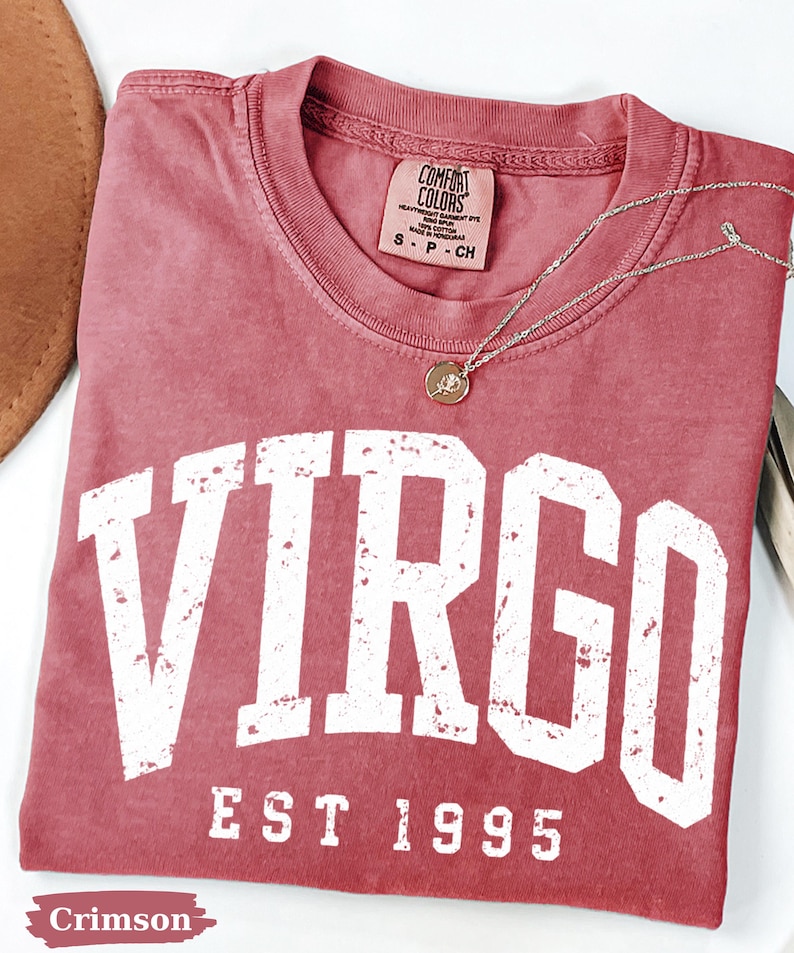 May include: A crimson short-sleeved t-shirt featuring the white distressed text VIRGO and EST 1995 printed on the front. The shirt has a Comfort Colors brand label at the inner neckline and is accompanied by a thin gold-toned chain necklace.