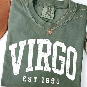May include: A moss green crew neck t-shirt featuring the white distressed text VIRGO and EST 1995 printed on the front. The shirt has a Comfort Colors label at the inner neckline indicating size S, and a thin gold chain necklace rests on the fabric.