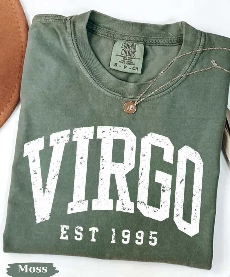 May include: A moss green crew neck t-shirt featuring the white distressed text VIRGO and EST 1995 printed on the front. The shirt has a Comfort Colors label at the inner neckline indicating size S, and a thin gold chain necklace rests on the fabric.