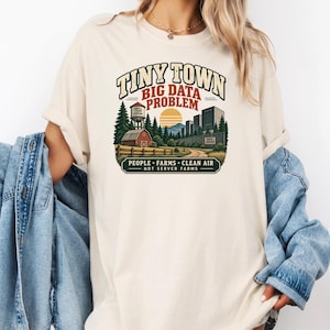 May include: A cream-colored t-shirt featuring a graphic print with the text "TINY TOWN BIG DATA PROBLEM" above an illustration of a barn, a water tower, and a data center. Below the image, the text reads "PEOPLE FARMS CLEAN AIR NOT SERVER FARMS."