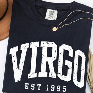 May include: A black short-sleeved t-shirt featuring the white distressed text VIRGO EST 1995 printed across the chest. The shirt has a crew neckline and a Comfort Colors brand label at the inner collar. A thin gold-toned chain necklace rests on top.
