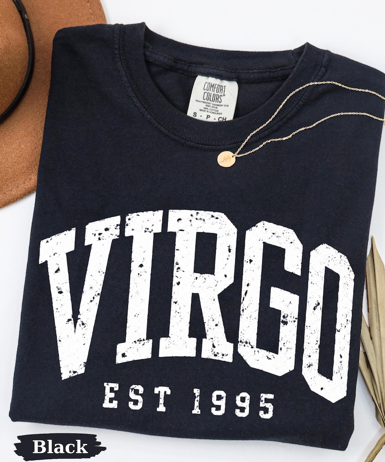 May include: A black short-sleeved t-shirt featuring the white distressed text VIRGO EST 1995 printed across the chest. The shirt has a crew neckline and a Comfort Colors brand label at the inner collar. A thin gold-toned chain necklace rests on top.