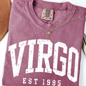 May include: A berry-colored short-sleeve t-shirt with a crew neckline. The shirt features the word VIRGO in large, distressed white block letters, with the text EST 1995 underneath. A thin gold-toned chain necklace with a small round pendant rests on the fabric.