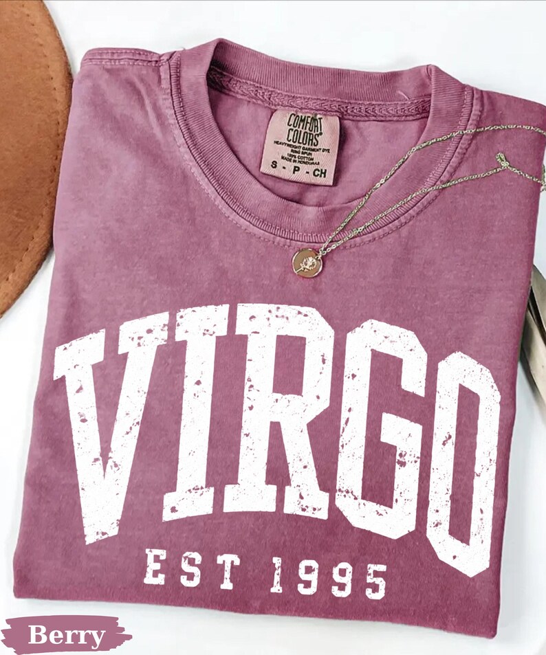 May include: A berry-colored short-sleeve t-shirt with a crew neckline. The shirt features the word VIRGO in large, distressed white block letters, with the text EST 1995 underneath. A thin gold-toned chain necklace with a small round pendant rests on the fabric.