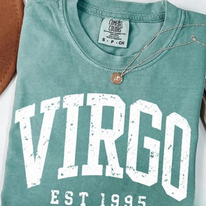 May include: A light green t-shirt featuring the word VIRGO in large, distressed white block letters above the text EST 1995. A thin silver chain necklace with a small circular pendant rests on the neckline, which has a Comfort Colors label.