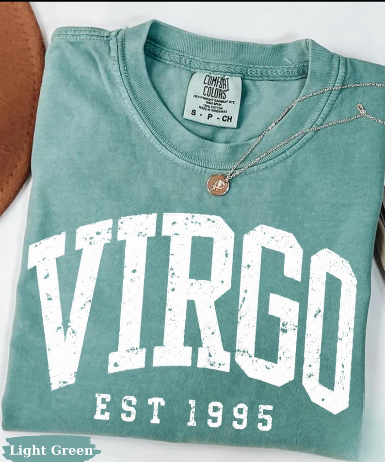 May include: A light green t-shirt featuring the word VIRGO in large, distressed white block letters above the text EST 1995. A thin silver chain necklace with a small circular pendant rests on the neckline, which has a Comfort Colors label.