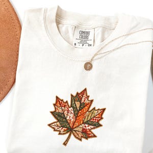 May include: An ivory t-shirt featuring a central embroidered maple leaf design composed of various patchwork-style fabric patterns in shades of orange, green, and brown. A thin gold-tone necklace with a small circular pendant rests on the neckline.