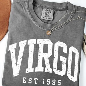 May include: A charcoal gray t-shirt with a distressed white graphic that reads VIRGO EST 1995. The shirt features a Comfort Colors brand label at the inner neckline, indicating size S, and is paired with a delicate gold-toned chain necklace.