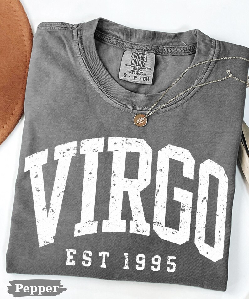 May include: A charcoal gray t-shirt with a distressed white graphic that reads VIRGO EST 1995. The shirt features a Comfort Colors brand label at the inner neckline, indicating size S, and is paired with a delicate gold-toned chain necklace.