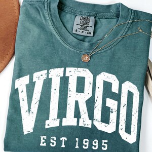 May include: A blue spruce colored t-shirt with a distressed white graphic that reads VIRGO EST 1995. The shirt features a crew neckline and a Comfort Colors brand label at the inner neck. A thin gold chain necklace with a small round pendant rests on top.