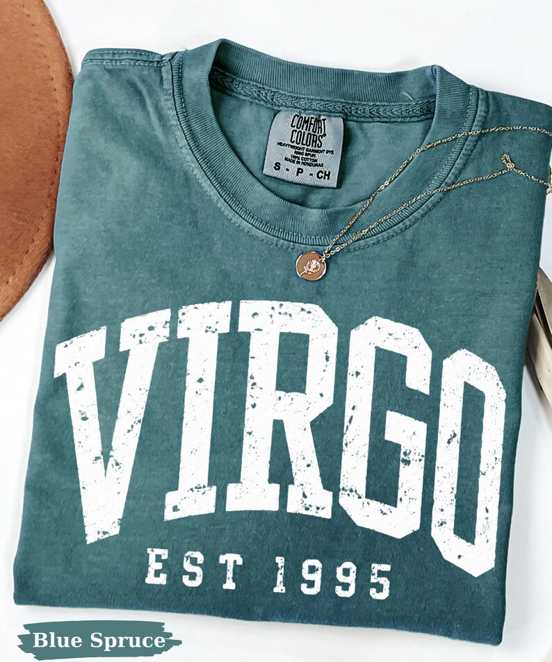 May include: A blue spruce colored t-shirt with a distressed white graphic that reads VIRGO EST 1995. The shirt features a crew neckline and a Comfort Colors brand label at the inner neck. A thin gold chain necklace with a small round pendant rests on top.