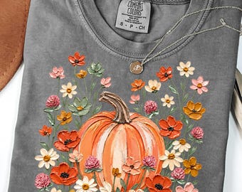 May include: A charcoal grey short-sleeved t-shirt featuring a central graphic of a large orange pumpkin surrounded by a variety of colourful painted-style flowers in shades of red, pink, orange, and white. The shirt has a Comfort Colors brand label.