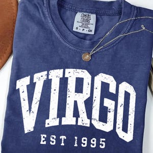 May include: A navy blue short-sleeve t-shirt with a distressed white graphic that reads VIRGO EST 1995. The shirt features a crew neckline and a Comfort Colors brand label on the inside collar. A thin gold-toned chain necklace with a small round pendant rests on the fabric.