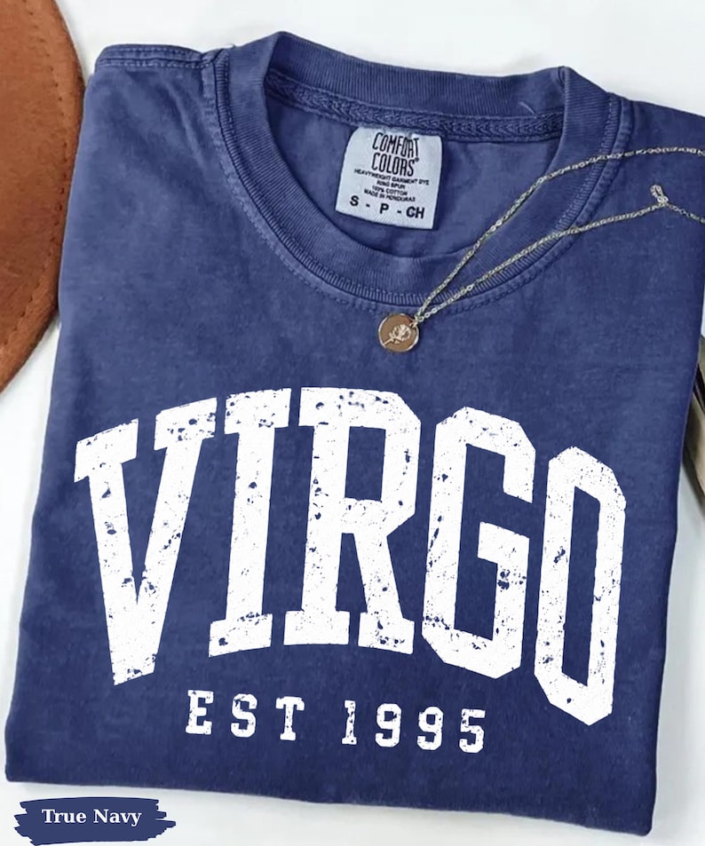 May include: A navy blue short-sleeve t-shirt with a distressed white graphic that reads VIRGO EST 1995. The shirt features a crew neckline and a Comfort Colors brand label on the inside collar. A thin gold-toned chain necklace with a small round pendant rests on the fabric.