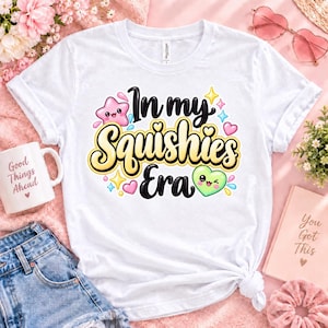 May include: A white short-sleeved t-shirt featuring a colorful graphic print that reads In my Squishies Era in bold black and yellow lettering, surrounded by small cartoon-style pink stars, green hearts, and various small sparkles.