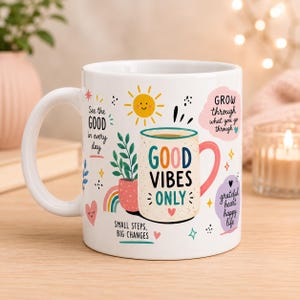 May include: White ceramic mug with a colorful design. The mug features the text "GOOD VIBES ONLY" and other positive affirmations. A smiling sun, rainbow, and potted plant are also included in the design. The mug has a pink handle.