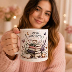 May include: A white ceramic mug featuring a stack of colorful books, a small teacup, a candle, and butterflies. The text "JUST ONE MORE CHAPTER" is printed in black above the books. The design includes floral accents and a crescent moon.