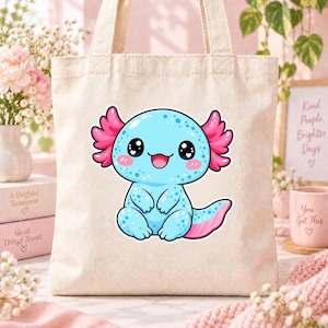 May include: A beige canvas tote bag featuring a centered graphic of a light blue axolotl with bright pink gills, large black eyes with white highlights, and a small smiling mouth. The axolotl has a spotted pattern and a pink-tipped tail.