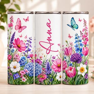 May include: Three cylindrical tumblers with a white background featuring a colorful floral pattern of pink, blue, purple, and white flowers with butterflies. The center tumbler displays the name Anna in a pink cursive script font.