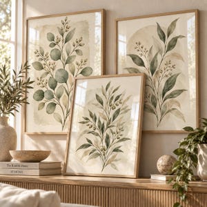 Peut inclure: Trois cadres muraux présentant des illustrations botaniques à l'aquarelle de branches d'eucalyptus et d'olivier vertes avec des baies sur fond crème, disposés dans des cadres en bois clair sur une surface en bois avec des livres et objets.