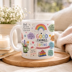 Puede incluir: Una taza de cerámica blanca con un asa grande, decorada con texto e ilustraciones coloridas. La taza presenta un arcoíris, flores y frases como "Good Things Are Coming". Está sobre una bandeja de madera.