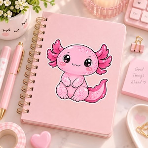 May include: A light pink spiral-bound notebook featuring a cartoon axolotl sticker on the cover. The axolotl is pink with dark pink gills, large black eyes with heart-shaped highlights, and a spotted body. The notebook is surrounded by pink stationery.