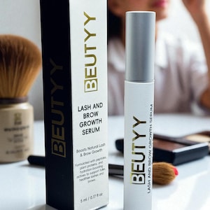 May include: A white and black box and a white tube of BEUTYY Lash and Brow Growth Serum. The box and tube have gold lettering. The box reads "LASH AND BROW GROWTH SERUM" and "5 ml / 0.17 fl oz". A makeup brush is in the background.