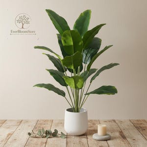 Puede incluir: Una planta verde artificial grande con hojas anchas en una maceta blanca. La planta está colocada sobre una superficie de madera, con una pequeña vela y una ramita decorativa cerca. El logotipo "EverBloomStore" es visible.