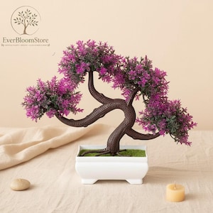 Könnte beinhalten: Ein Miniatur-Bonsai-Baum mit leuchtend lila Blüten und dunkelbraunen Ästen. Die künstliche Pflanze steht in einem weißen, rechteckigen Keramiktopf mit grüner Moosbasis. Das EverBloomStore-Logo befindet sich oben links.
