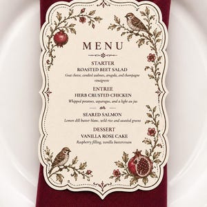 May include: An ornate menu card featuring a floral design, birds, and pomegranates. The menu details a starter of roasted beet salad, an entree of herb crusted chicken or seared salmon, and vanilla rose cake for dessert. It rests on a burgundy napkin.