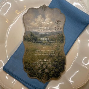 May include: A scalloped-edge menu card featuring a landscape illustration of a field and sky. The text reads Menu, Starter: Fresh Fig & Burrata Salad, Entree: Herb Grilled Filet Mignon or Lemon Thyme Chicken, and Dessert: Vanilla Bean Creme Brulee.